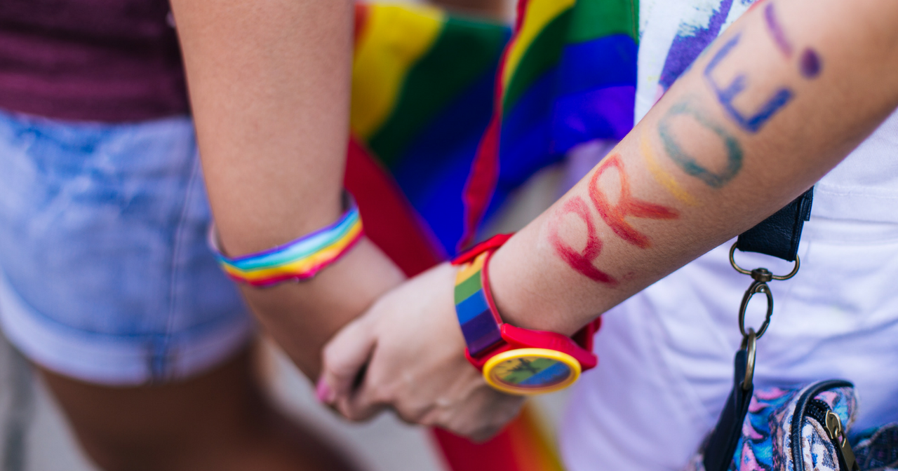 To people holding hands with rainbow accessories and the word PRIDE written on one forearm