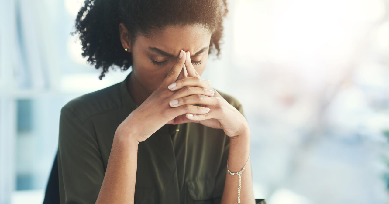 A Black woman sits with her hands over part of her face looking stressed. 