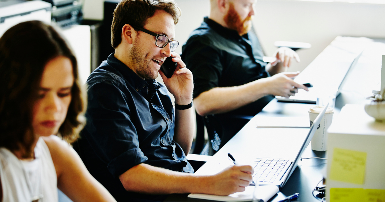 Three people sit at their computers while talking on the phone in an office. 