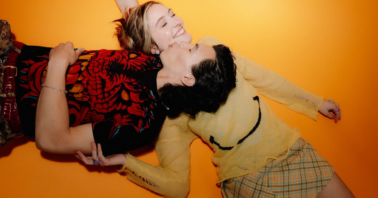A teen couple lays on the ground and laughs. The man kisses the woman's cheek as his head rests on her chest. 