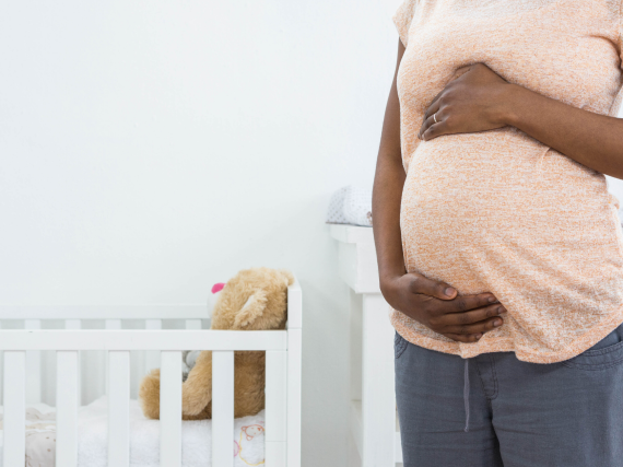 A pregnant Black woman holding her belly while standing next to a crib. 