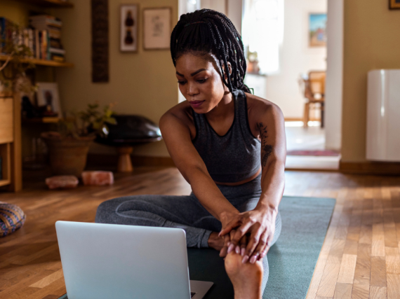 A woman stretches on a yoga mat while looking at her computer. 