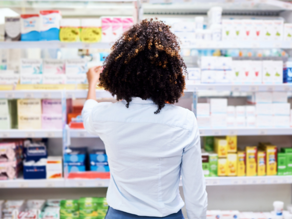 A young person stands with their back to the photo and considers a row of period products in a pharmacy. 