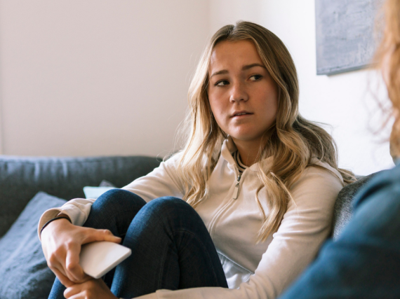 A concerned looking teen hugs her knees to her chest while talking to an adult. 