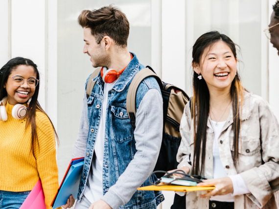 A group of four smiling teens walk together with their bookbags. 