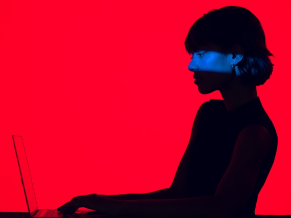 A woman in shadow types on a laptop. The background is bright red.
