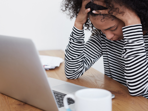A woman sits at a computer, stressed, with her hands on her head. 