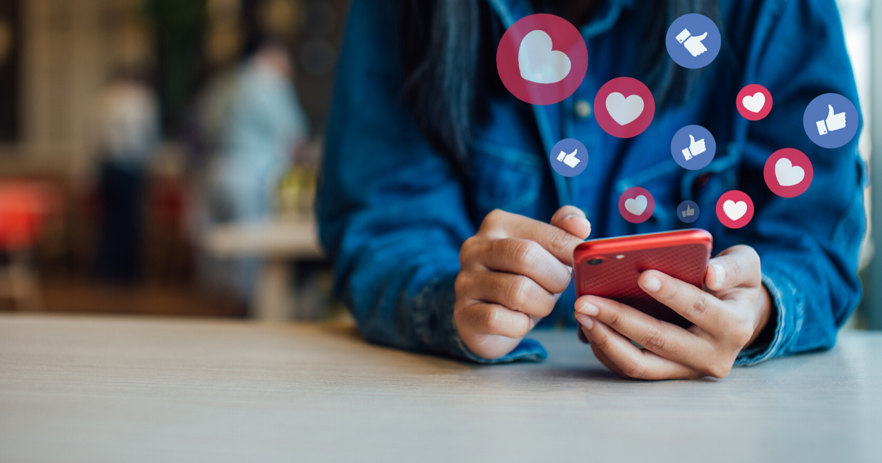 A pair of hands hold a phone with social media icons floating above it. 